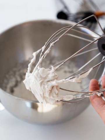 A wire whisk with coconut whipped cream clinging to it over a silver mixing bowl filled with whipped cream.