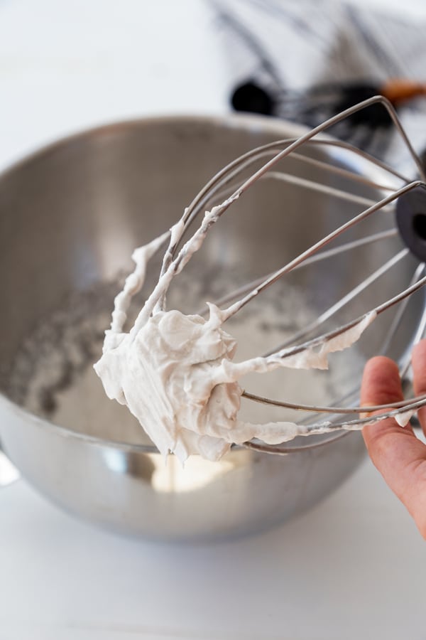 A wire whisk with coconut whipped cream clinging to it over a silver mixing bowl filled with whipped cream.