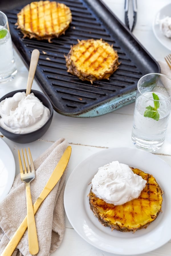 A grill pan with grilled pineapple and a white plate with a slice of grilled pineapple and whipped cream and a black bowl with cream and gold utensils. 