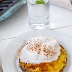 A slice of grilled pineapple with whipped cream and cinnamon on a white plate with a glass of water in the background.