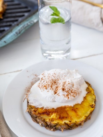 A slice of grilled pineapple with whipped cream and cinnamon on a white plate with a glass of water in the background.