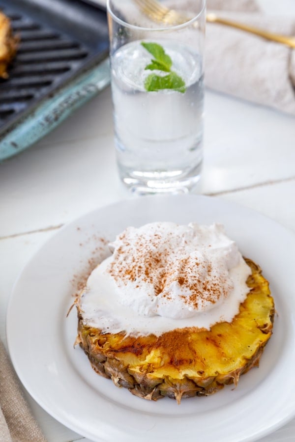 A slice of grilled pineapple with whipped cream and cinnamon on a white plate with a glass of water in the background.