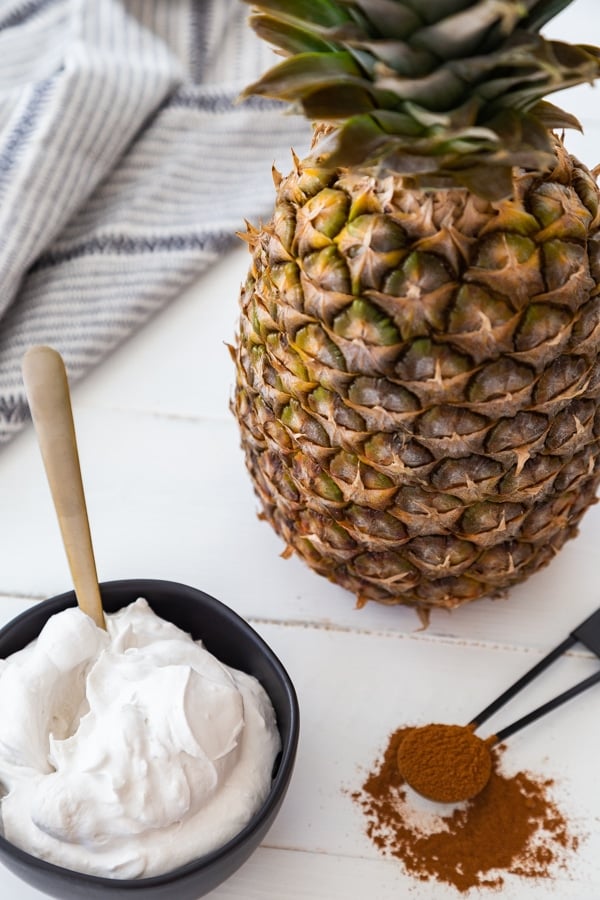 A black bowl filled with whipped cream and a gold spoon in the bowl, and a black measuring spoon with cinnamon spilling off of it and a pineapple on a white wood table. 