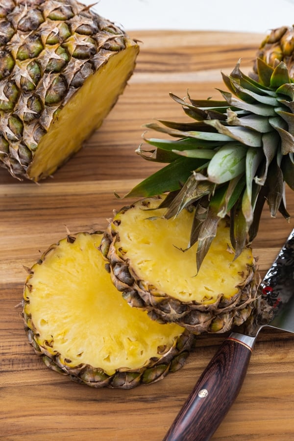 2 slices of pineapple with a knife on a wood board and the crown of the pineapple leaning on one slice an the rest of the whole pineapple in the background.