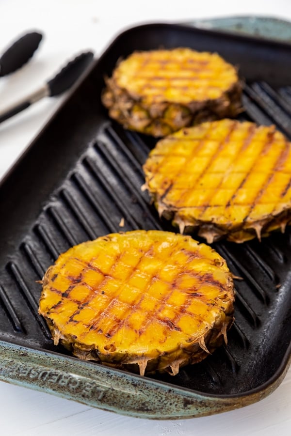 Three slices of grilled pineapple on an iron grill pan with tongs in the background. 