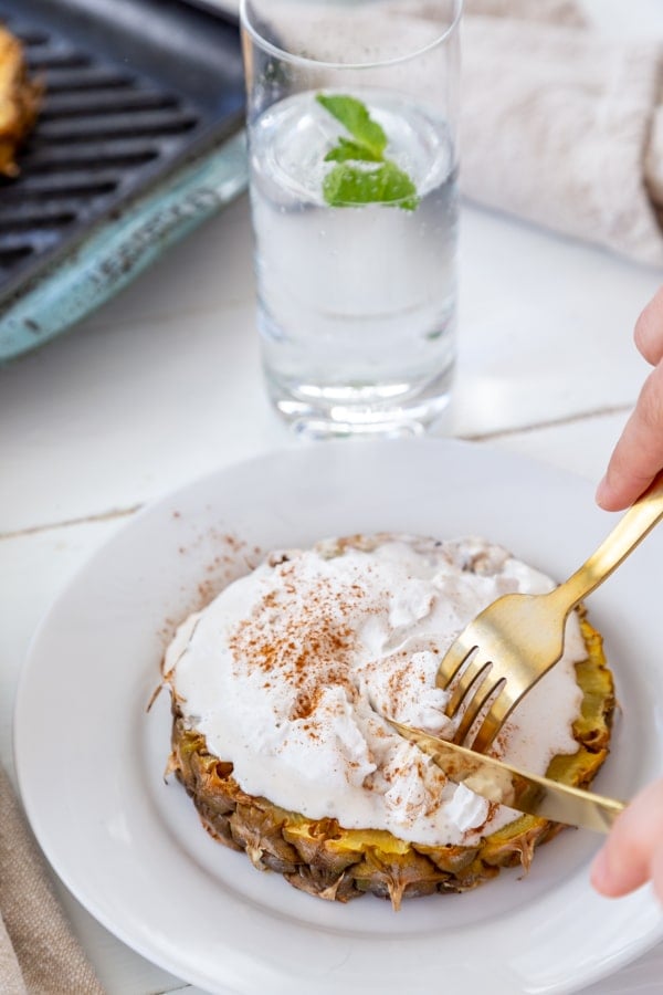 A slice of grilled pineapple with whipped cream and cinnamon on a white plate with hands holding a gold fork and knife, cutting into it. 