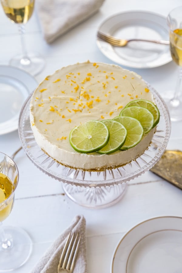 A cheesecake on a glass cake plate with lime slices and lime and orange zest on a white table with plates and glasses of wine next to it. 