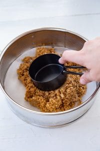 vegan cheesecake crust being pressed down into springform pan lined with parchment paper by a black measuring cup and hand on a white board