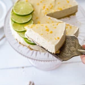 A cheesecake with lime slices on a glass cake stand with a silver cake server holding a slice of pie over the whole cake.