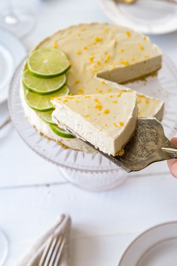 A cheesecake with lime slices on a glass cake stand with a silver cake server holding a slice of pie over the whole cake.