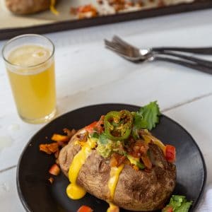 A stuffed potato on a black plate with a glass of beer and utensils, and a a platter with stuffed potatoes in the background.