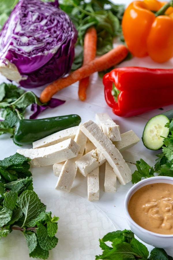 Spinach and mint leaves, basil, bell peppers, red cabbage, tofu strips, and a white bowl with peanut sauce on a white table. 