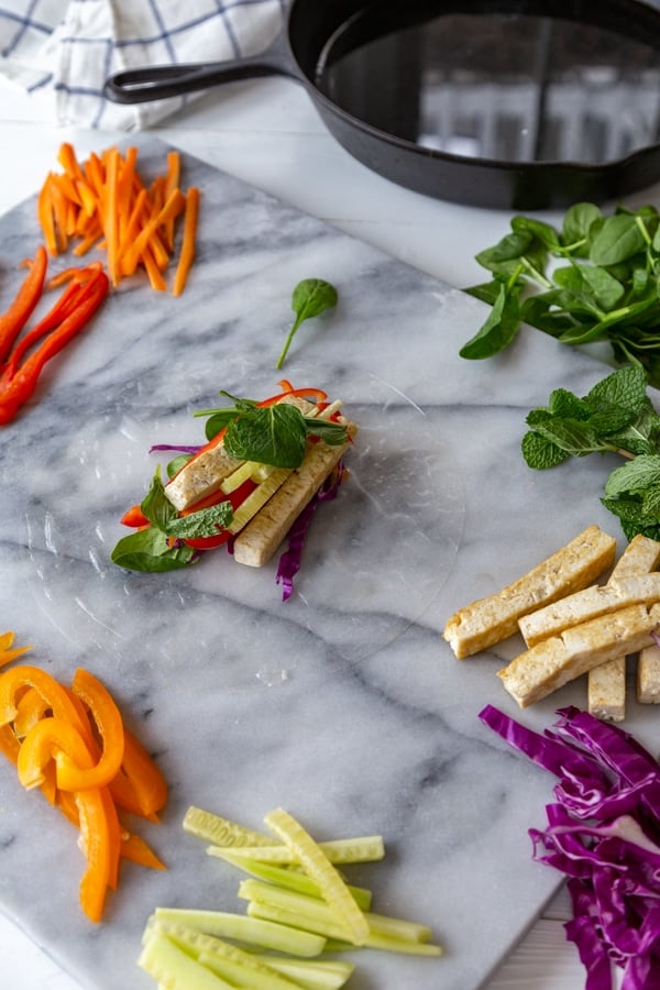 A spring roll wrapper on a marble board with vegetables and greens on top.