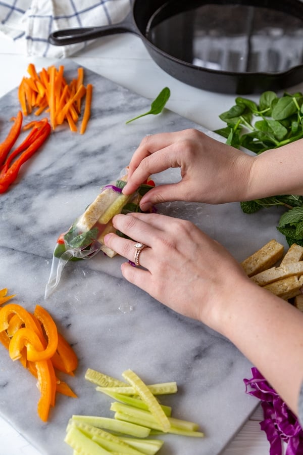 Two hands rolling up a tofu and veggie spring roll on a marble board.