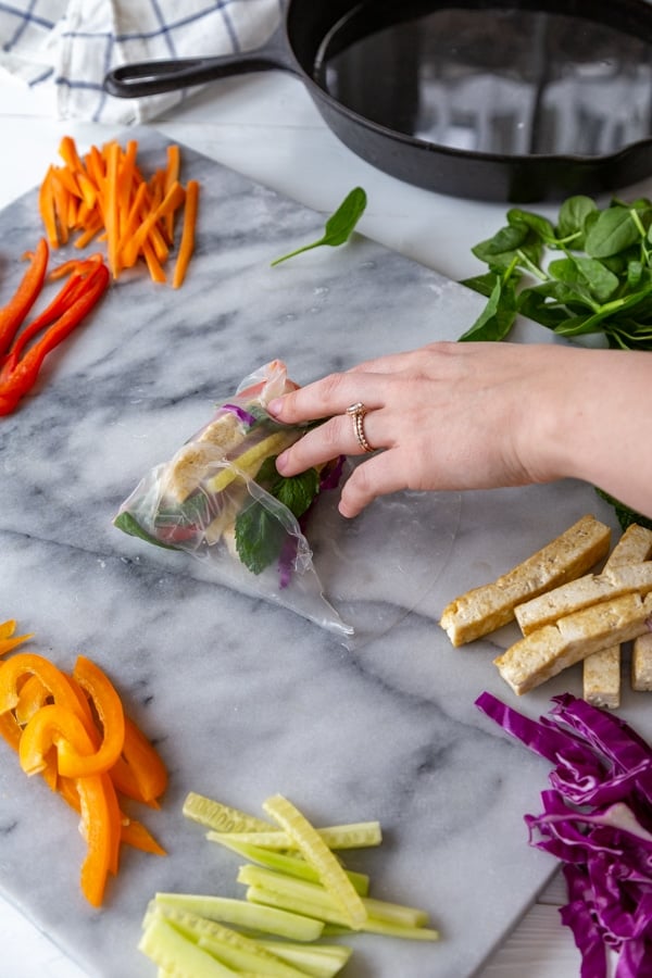 A hand rolling a spring roll with veggies on a marble board.