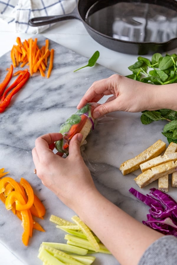 Two hands rolling a spring roll with vegetables on a marble board with the ingredients next to it.
