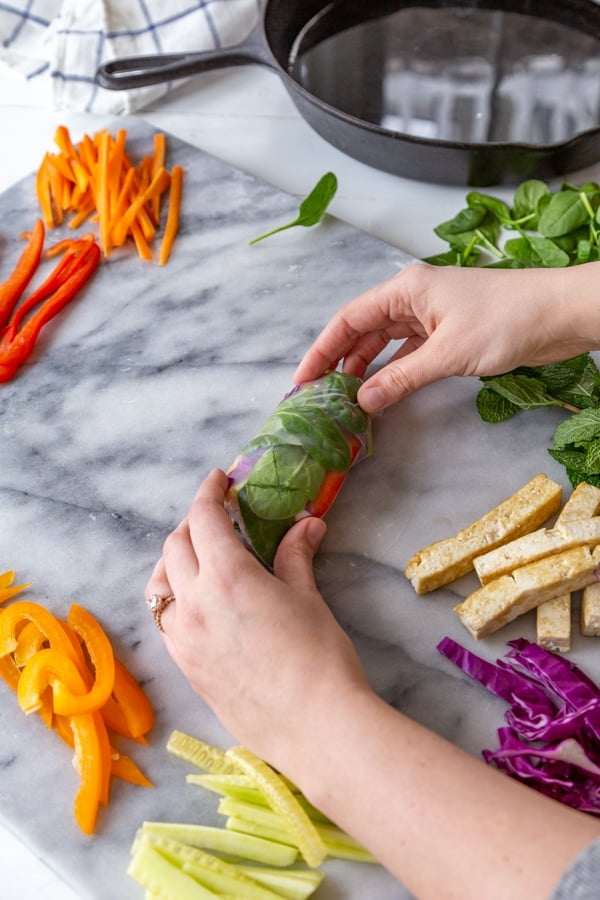 Two hands rolling a veggie spring roll on a marble board with veggies and an iron skillet with water next to it.