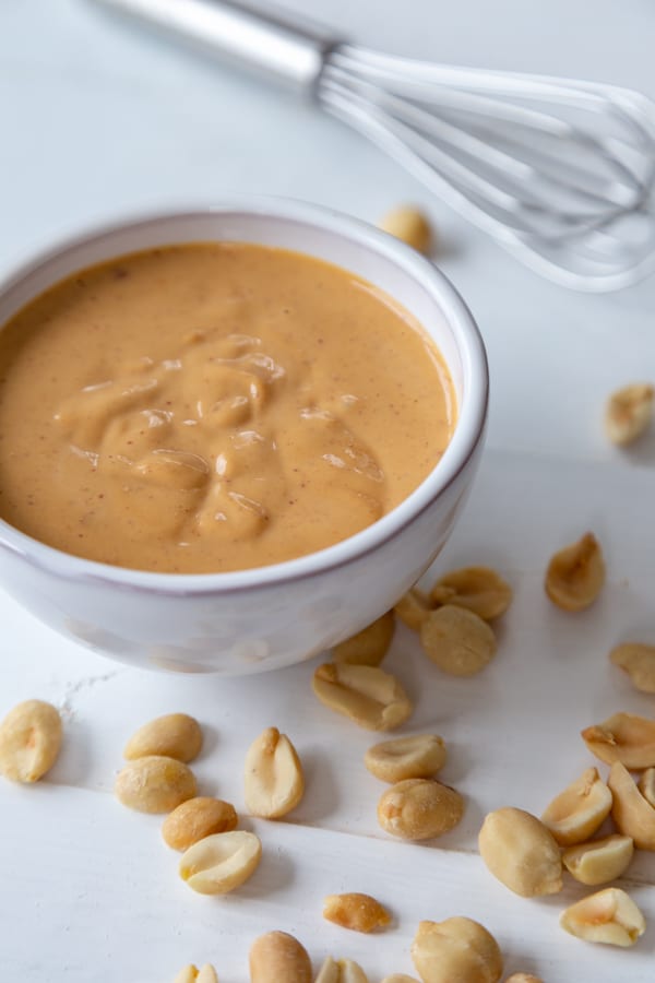 A white bowl with peanut sauce and peanuts and a silver whisk next to it on a white wood table. 