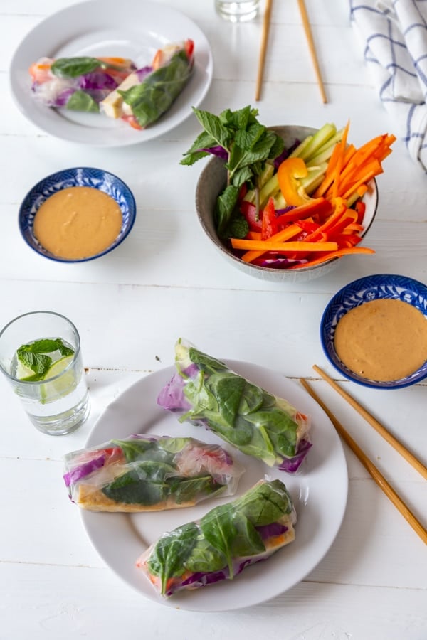 A white table with two white plates with spring rolls and two blue bowls of peanut sauce, a glass of water, and chopsticks.