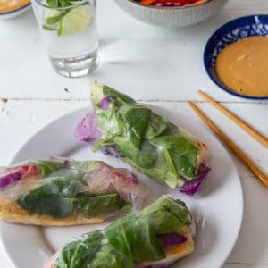 A white plate with three spring rolls and a blue bowl with peanut sauce, a glass of water, a bowl of veggies, and chopsticks on a white table.