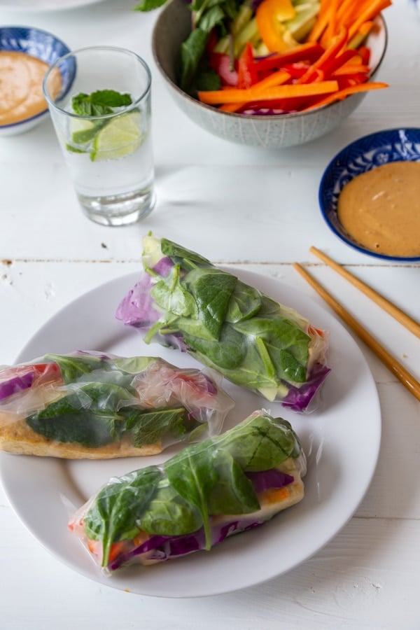 A white plate with three spring rolls and a blue bowl with peanut sauce, a glass of water, a bowl of veggies, and chopsticks on a white table. 