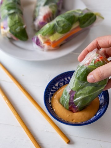 A hand dipping a spring roll into a blue and white dish of peanut sauce with a plate of spring rolls and chopsticks in the background.