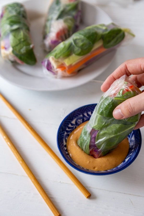 A hand dipping a spring roll into a blue and white dish of peanut sauce with a plate of spring rolls and chopsticks in the background.