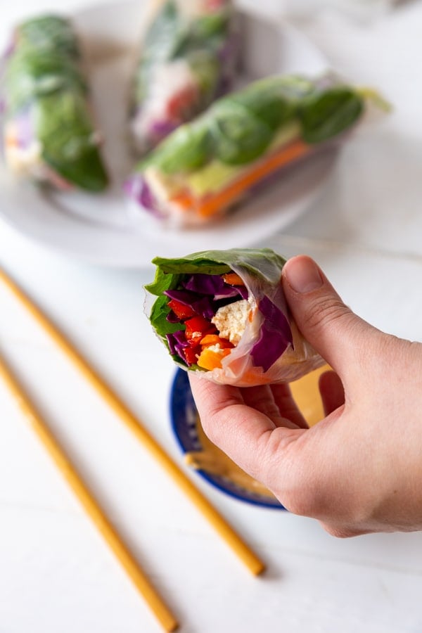 A hand holding a veggie spring roll and a white plate with 3 rolls and chopsticks on a white table. 