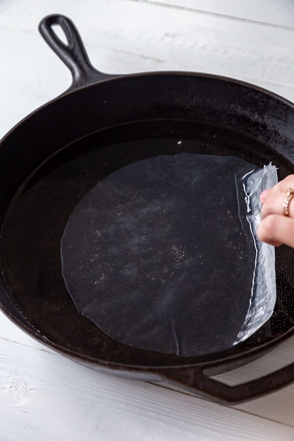 A piece of rice paper in water in a black skillet with a hand pulling the paper out of the pan. 