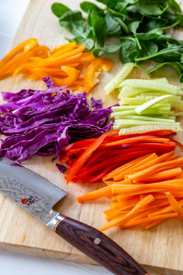 A cutting board and a knife with sliced bell peppers, red cabbage, cucumber, and carrots.