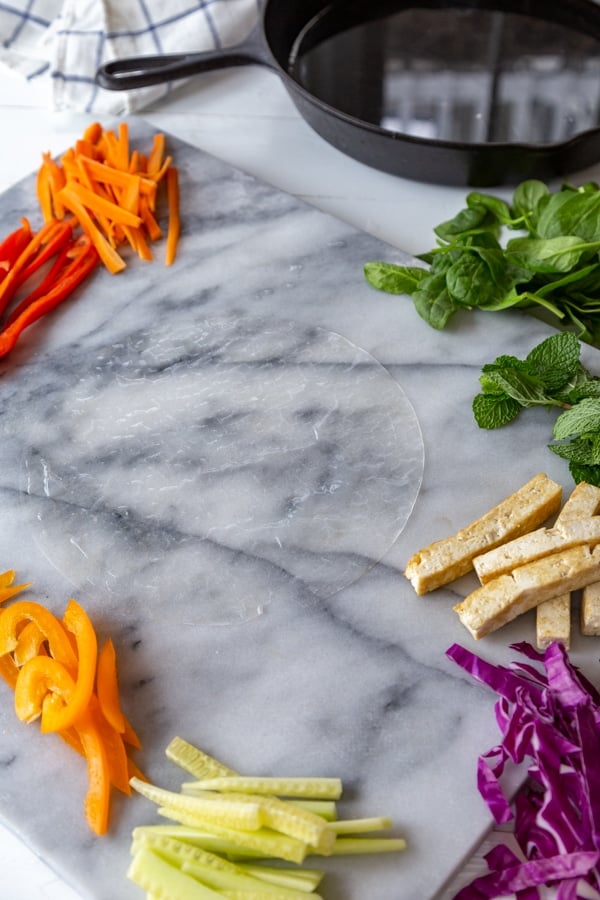 Rice paper and sliced vegetables on a marble slab with an iron skillet in the background.