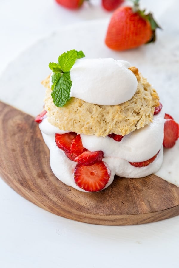 Strawberry shortcake on a marble and wood board with a sprig of mint on top and strawberries in the background. 