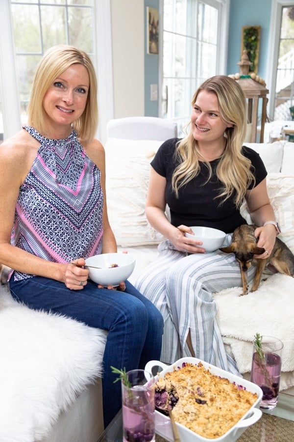 Two blonde women on a white couch holding bowls of blueberry crumble and a dog is next to one of the women.