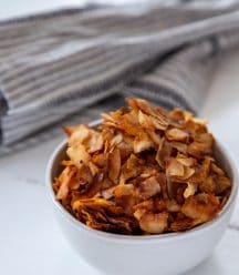 A white bowl filled with coconut bacon on a white table with a gray towel next to it.