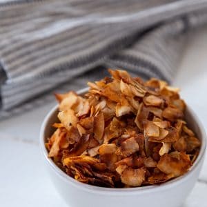 A white bowl filled with coconut bacon on a white table with a gray towel next to it.