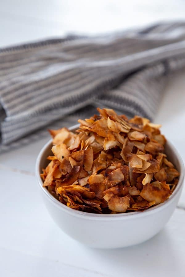 A white bowl filled with coconut bacon on a white table with a gray towel next to it.