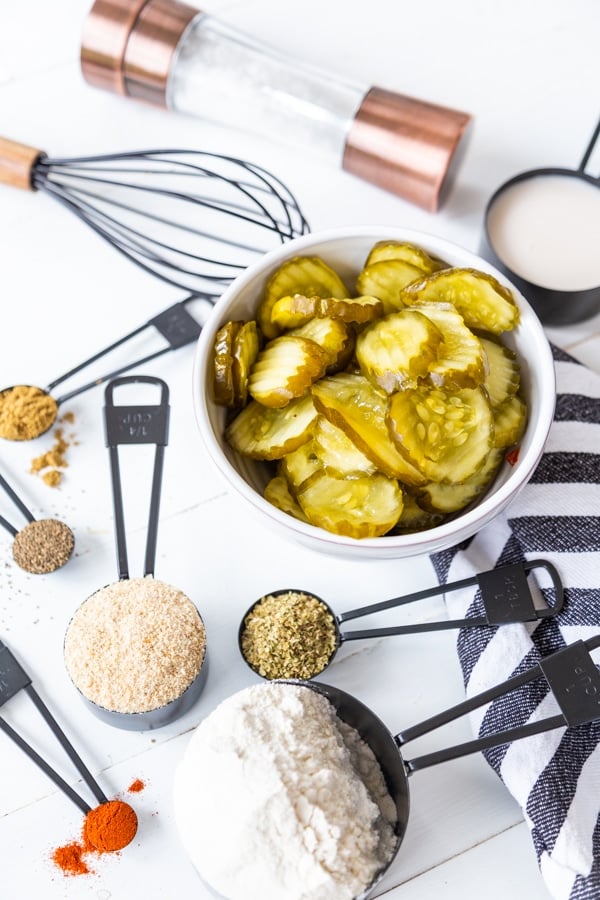 A bowl of pickle chips and black measuring spoons with flour and spices on a blue and white striped towel.