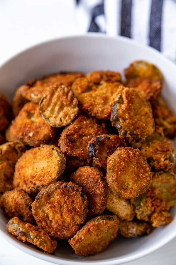 A close up shot of fried pickles in a white bowl with a striped towel next to the bowl.