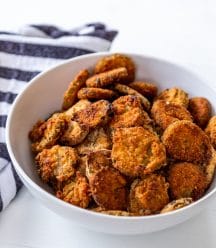 A white bowl filled with fried pickles on a white table with a blue and white stripe towel next to the bowl.
