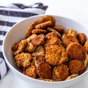 A white bowl filled with fried pickles on a white table with a blue and white stripe towel next to the bowl.