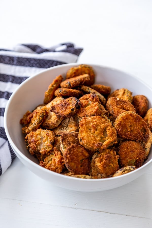 A white bowl filled with fried pickles on a white table with a blue and white stripe towel next to the bowl.