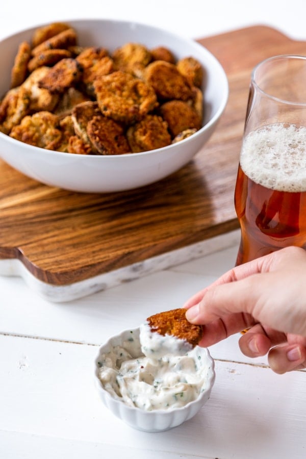 A hand dipping a fried pickle into ranch dressing with a bowl of fried pickles and a glass of beer in the background.