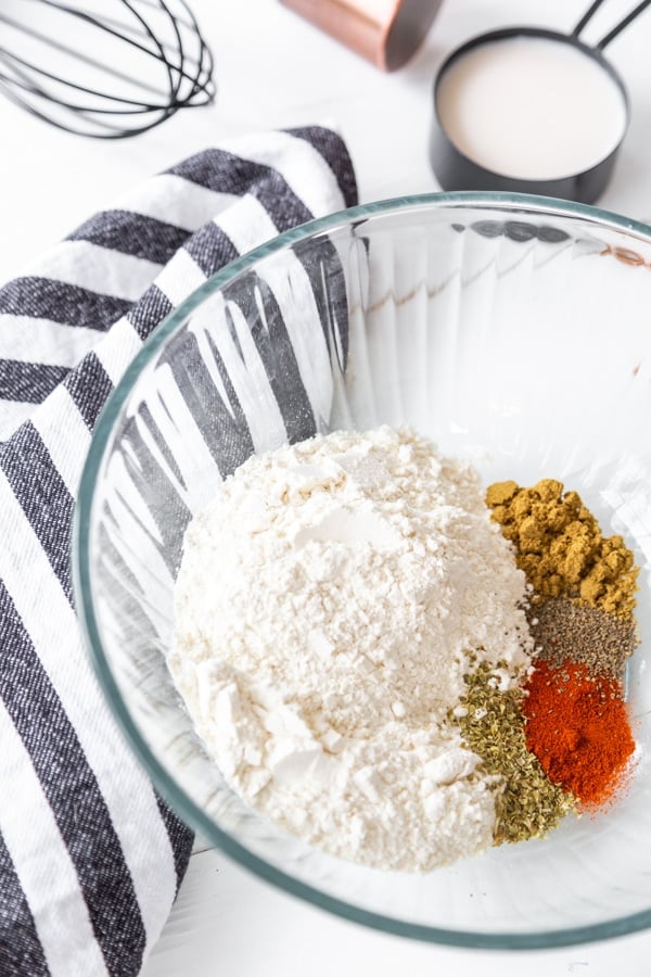 A glass bowl with flour and spices and a striped towel next to the bowl and a whisk and measuring cup with milk behind it.