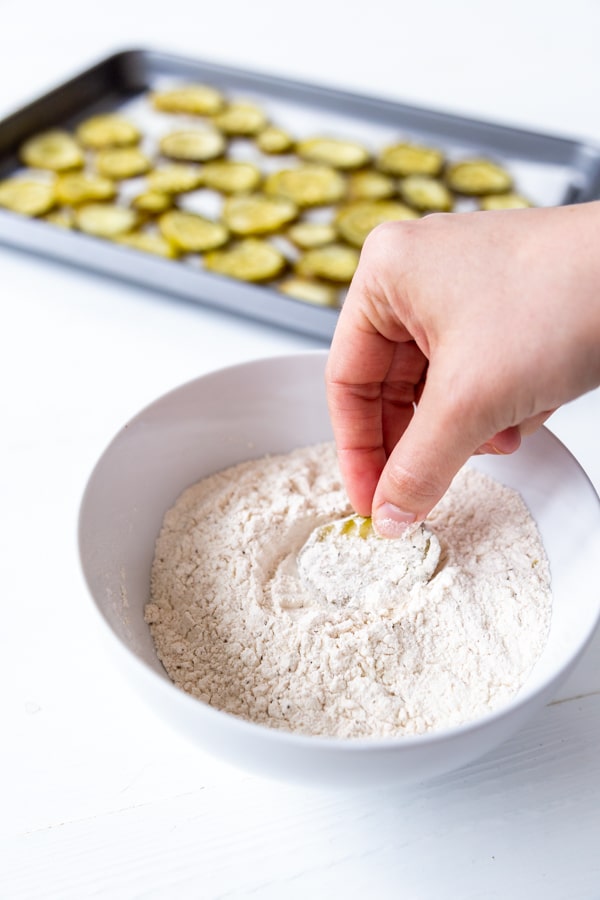 A hand dipping a pickle chip into a bowl of flour with a pan of pickle chips in the background