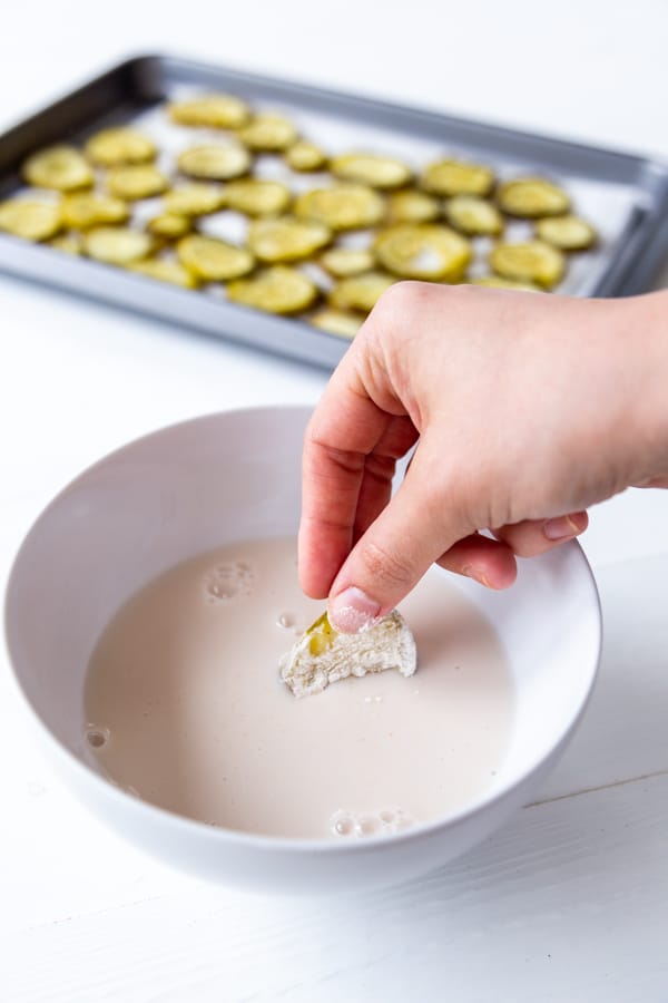 A hand dipping a flour covered pickle chip in a bowl of milk with a tray of pickle chips in the background.