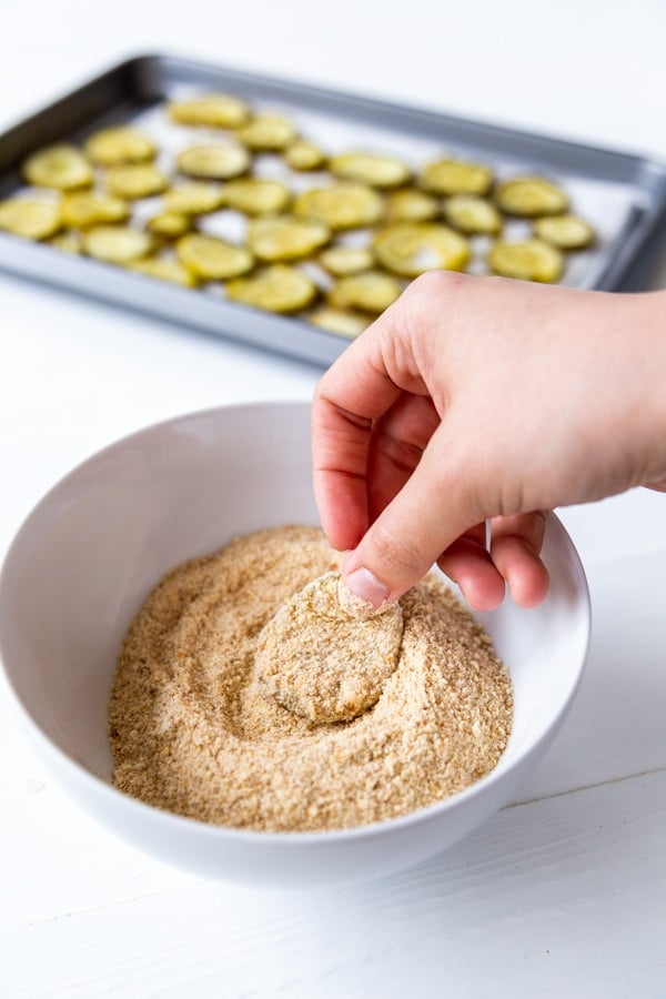 A hand dipping a flour and milk covered pickle chip in a bowl of bread crumbs with a pan of pickle chips in the background.