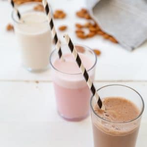 A glass of chocolate, strawberry, and plain almond milk in a row with black and white straws in the glasses and almonds scattered next to them on a white table.