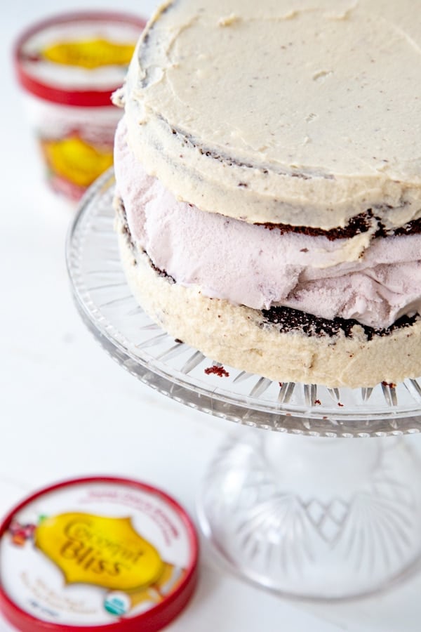 A chocolate and cherry ice cream cake on a cake stand with 2 pints of ice cream next to it.