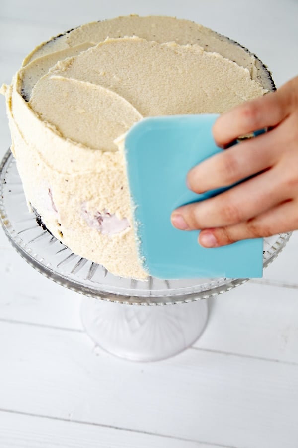 A hand spreading vanilla frosting on a cake with a blue cake scraper.