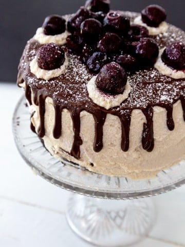 A cake on a clear glass cake stand with white frosting and a chocolate ganache dripping down the sides, and cherries on top.
